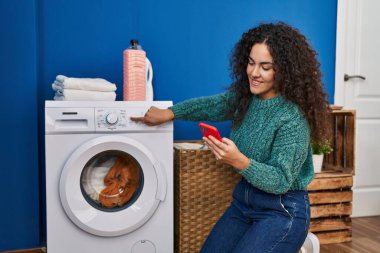 Çamaşır odasında çamaşır makinesini çalıştırmak için akıllı telefon kullanan güzel İspanyol kadın.