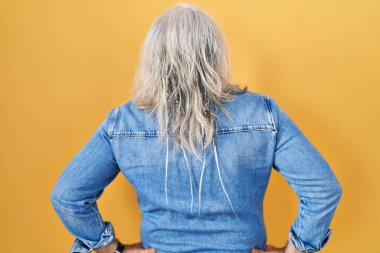 Sarı arka planda gri saçlı, orta yaşlı bir adam kolları vücudunda geriye doğru bakıyor. 