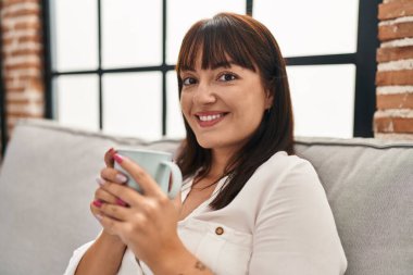 Genç ve güzel İspanyol kadın evdeki kanepede oturup kahve içiyor.