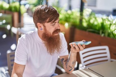 Young redhead man talking on the smartphone sitting on table at coffee shop terrace