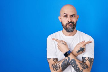 Mavi arka planda dikilen İspanyol dövmeli adam her iki tarafı da parmaklarıyla işaret ediyor, farklı yönlere. 