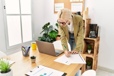 Genç sarışın iş kadını ofisteki evraklarda dizüstü bilgisayar kullanıyor.