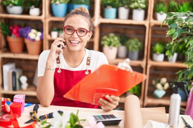 Young beautiful hispanic woman florist talking on smartphone reading clipboard at florist