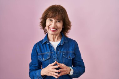 Orta yaşlı bir kadın pembe arka planda el ele tutuşup gülümseyerek gevşemiş ve neşeli gülümsüyor. Başarı ve iyimserlik 