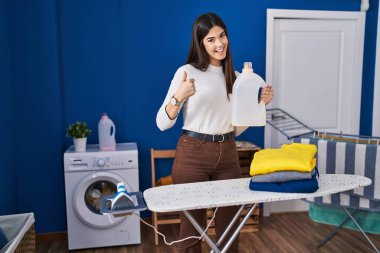 Ütü yaptıktan sonra katlanmış çamaşırları olan genç esmer kadın mutlu ve pozitif gülümsüyor, başparmağıyla mükemmel ve onay işareti yapıyor. 