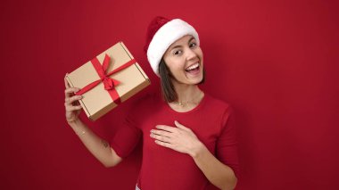 Genç, güzel, İspanyol bir kadın Noel şapkası takıyor ve kırmızı arka planda hediyeler tutuyor.