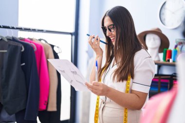 Young beautiful hispanic woman tailor talking on smartphone looking clothing design at clothing factory