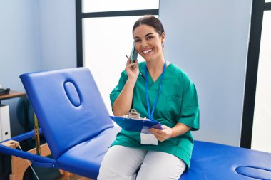 Young beautiful hispanic woman physiotherapist reading document talking on smartphone at rehab clinic