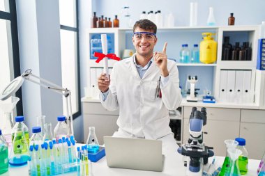 Laboratuvarda çalışan Latin asıllı genç bir adam elinde diplomayla mutlu mesut gülümsüyor. 