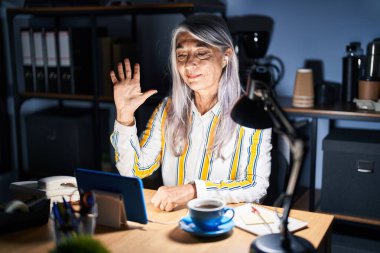 Gri saçlı orta yaşlı bir kadın gece ofiste çalışıyor. Beş numaralı parmaklarıyla işaret ediyor ve gülümsüyor. Kendinden emin ve mutlu.. 