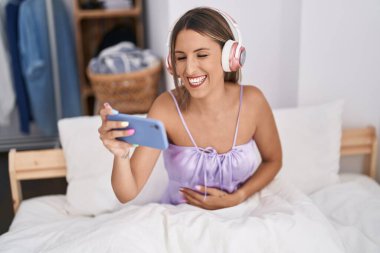 Young beautiful hispanic woman watching video on smartphone sitting on bed at bedroom