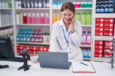 Genç sarışın eczacı, eczanede bilgisayarla akıllı telefondan konuşuyor.