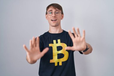 Bitcoin t-shirt giyen beyaz sarışın adam korku ifadesinden korkuyor ve dehşete düşüyor. El hareketlerini durdur, şok içinde bağır. panik konsepti. 