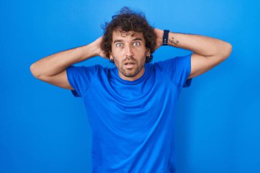 Mavi arka planda dikilen İspanyol genç adam deli ve elleri başının üstünde korkmuş, korkmuş ve ağzı açık şoktan şaşırmış. 