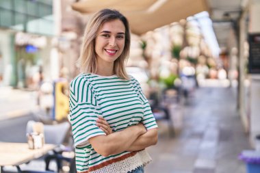 Kendine güvenen genç bir kadın caddede kollarını kavuşturmuş duruyor.