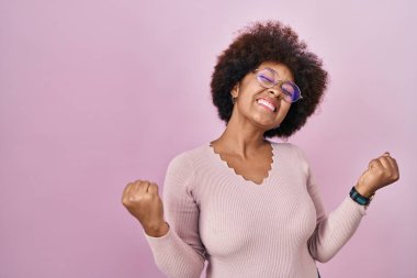 Pembe arka planda duran genç Afro-Amerikalı kadın kolları havada ve gözleri kapalı bir şekilde başarının şaşkınlığını ve şaşkınlığını yaşıyor. Kazanan konsept. 