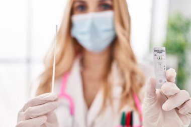 Young blonde woman doctor wearing medical mask holding covid antigen test at clinic