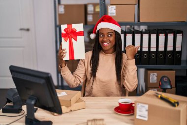 Saçları örgülü genç bir Afrikalı. Noel 'de küçük bir iş yerinde çalışıyor. Başparmağını yana uzatıyor ve ağzı açık gülümsüyor. 