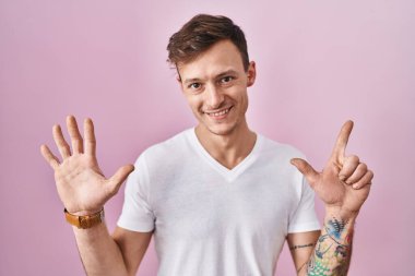 Pembe arka planda duran beyaz bir adam yedinci parmaklarını gösterip işaret ederken kendinden emin ve mutlu bir şekilde gülümsüyor.. 