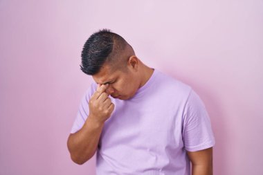 Young hispanic man standing over pink background tired rubbing nose and eyes feeling fatigue and headache. stress and frustration concept. 