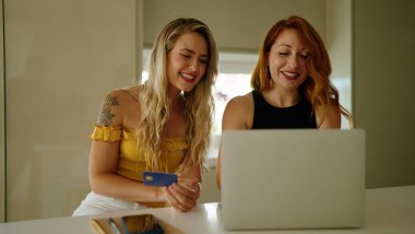 Two women using laptop and credit card sitting on table at kitchen