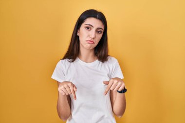 Sarı arka plan üzerinde duran genç ve güzel bir kadın üzgün ve üzgün görünüyor, parmaklarıyla yön gösteriyor, mutsuz ve depresif.. 