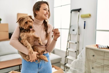 Young beautiful hispanic woman hugging dog holding key at new home