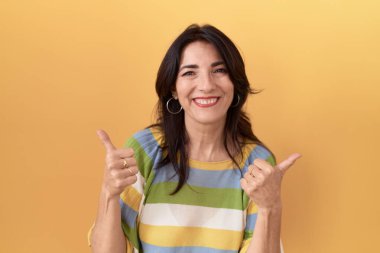 Orta yaşlı İspanyol kadın sarı arka plan başarı tabelasının üzerinde duruyor. Elleriyle olumlu jest yapıyor. Gülümsüyor ve mutlu. Neşeli ifade ve kazanan jest. 