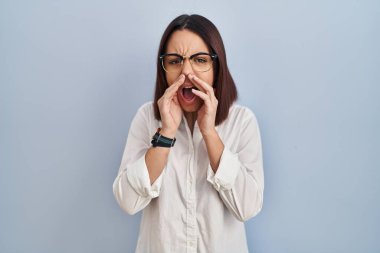 Young hispanic woman standing over white background shouting angry out loud with hands over mouth 