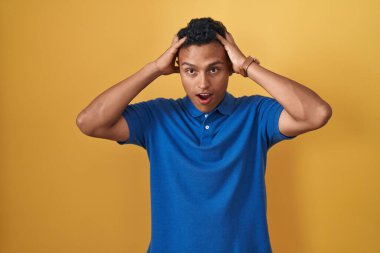 Sarı arka planda dikilen İspanyol genç adam deli ve elleri başının üstünde korkmuş, korkmuş ve ağzı açık şoktan şaşırmış. 