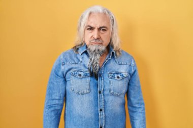 Sarı arka planda duran gri saçlı, kuşkucu ve gergin orta yaşlı bir adam sorun yüzünden üzgün. negatif kişi. 