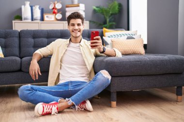 Genç İspanyol adam akıllı telefon kullanıyor. Evde yerde oturuyor.