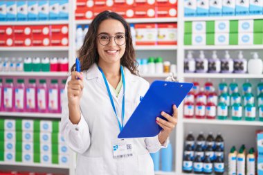 Young beautiful hispanic woman pharmacist holding clipboard and pen at pharmacy