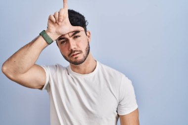 Hispanic man with beard standing over white background making fun of people with fingers on forehead doing loser gesture mocking and insulting. 