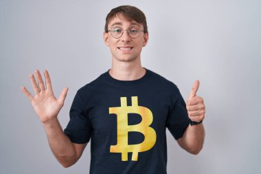 Beyaz, sarışın, bitcoin tişörtlü bir adam. Altıncı parmaklarını gösterip gösteriyor. Kendinden emin ve mutlu bir şekilde gülümsüyor.. 