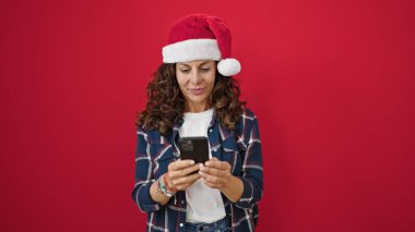 Orta yaşlı İspanyol kadın, izole edilmiş kırmızı arka plan üzerinde akıllı telefon kullanarak Noel şapkası takıyor.