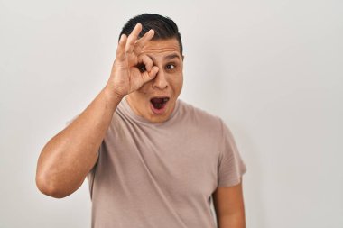 Beyaz arka planda dikilen Latin asıllı genç adam şaşırmış yüz ifadesiyle şaşırmış, parmaklarından bakarak hareket ediyor. İnançsız ifade. 