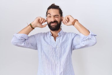 Hispanic man with beard wearing casual shirt covering ears with fingers with annoyed expression for the noise of loud music. deaf concept. 