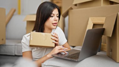 Genç beyaz kadın yeni evinde dizüstü bilgisayar paketi kullanıyor.