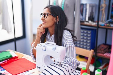 Young beautiful hispanic woman tailor smiling confident sitting on table at clothing factory