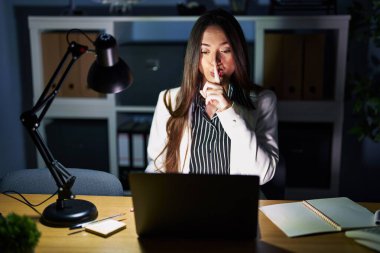 Genç esmer kadın geceleri ofiste laptopuyla çalışıyor ve parmakları dudaklarında sessiz olmak istiyor. sessizlik ve gizli konsept. 