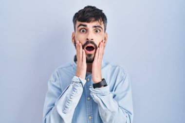 Mavi arka planda dikilen sakallı genç İspanyol adam korkmuş ve şok olmuş, şaşırmış ve şaşırmış bir yüz ifadesi var. 