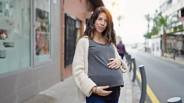 Young pregnant woman smiling confident touching belly at street