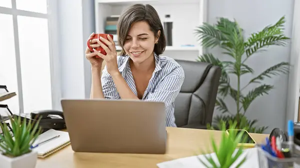 Confident young hispanic woman boss, with beautiful smile and short hair, happily working early morning at her office desk, drinking coffee while using her laptop for online business success.
