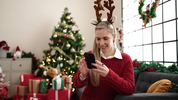 若いですブロンド女性使用してスマートフォン座っています上のソファによってクリスマスツリーホーム — ストック写真