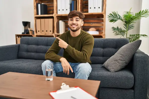 Arab man with beard working on depression at therapy office smiling cheerful pointing with hand and finger up to the side