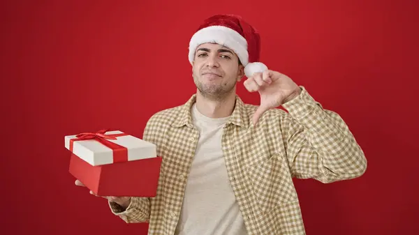 クリスマスの帽子をかぶった若いヒスパニック男性は 孤立した赤い背景の上に親指で贈り物を解放します — ストック写真
