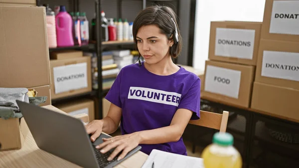 Heartwarming portrait of serious young beautiful hispanic female volunteer with short hair, sitting indoors at charity center room, using laptop and headphones while working online.