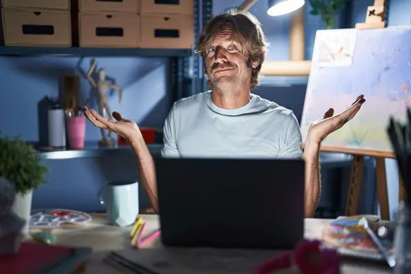 中年男子坐在艺术演播室里 晚上拿着笔记本电脑 毫无头绪 满脸困惑 手臂和双手高举 怀疑概念 — 图库照片
