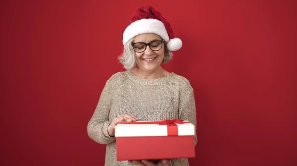 単離された赤い背景にクリスマスの帽子の荷を下すギフトを身に着けている灰色の髪を持つ中年の女性 — ストック写真